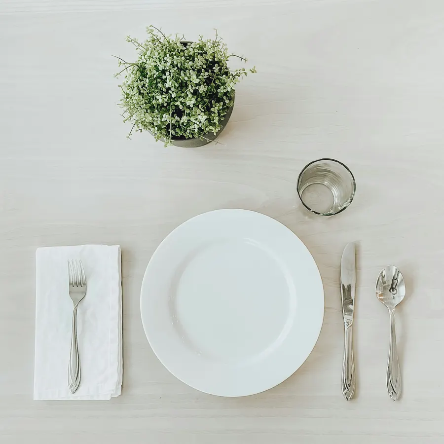 A single place setting from above: white plate, folded napkin, silver cutlery, a small vase of greenery.