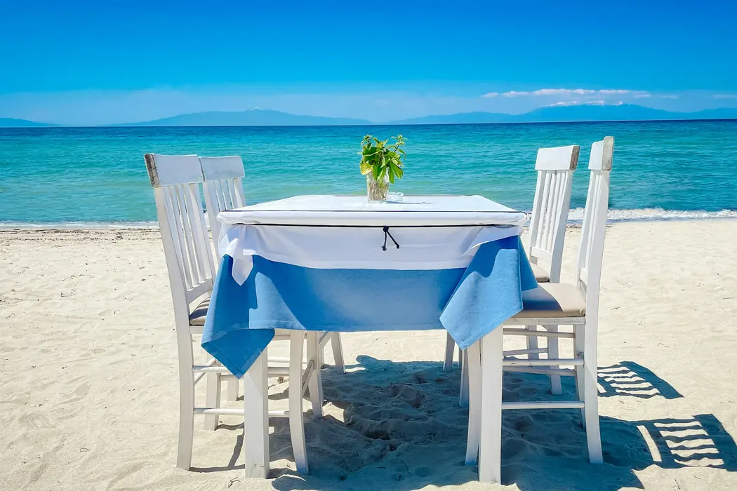 A white table dressed in blue linen set on the sand, looking straight out to turquoise water.
