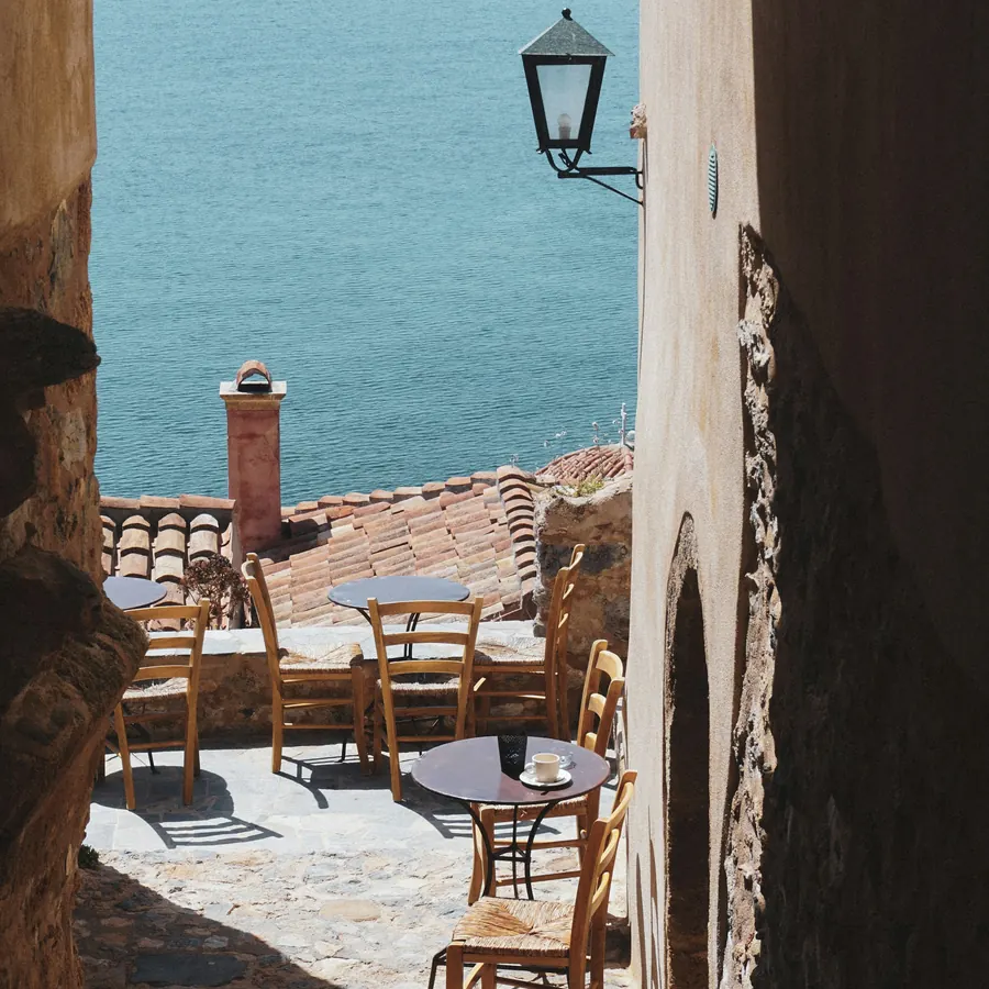 Tables on a stone terrace cut into the rock above a calm blue bay.